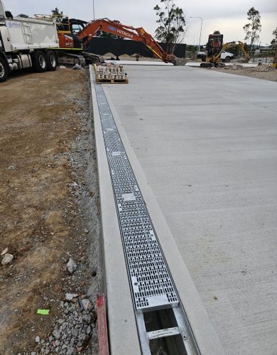 The construction site with a newly poured concrete slab, an integrated drainage system, and construction equipment, including a Hitachi excavator and a Supreme-branded truck, in use.