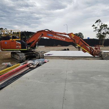 penrith christian school - a Hitachi excavator parked on a dirt surface next to a newly poured concrete slab, surrounded by construction materials and an overcast sky in the background.