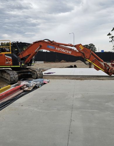 penrith christian school - a Hitachi excavator parked on a dirt surface next to a newly poured concrete slab, surrounded by construction materials and an overcast sky in the background.