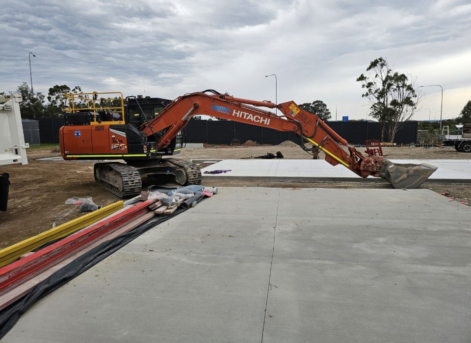 penrith christian school - a Hitachi excavator parked on a dirt surface next to a newly poured concrete slab, surrounded by construction materials and an overcast sky in the background.