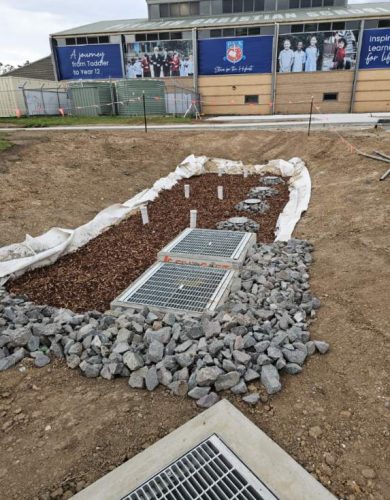 a landscaped drainage area with grated covers, gravel, and mulch surrounded by soil, situated near a building with signage promoting Penrith Christian School.